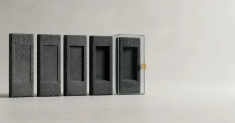 Five identical grey stone slabs in a row, each carved with the same blank panel but cut progressively deeper; the deepest-carved slab is sealed inside a glass case with a small brass clasp