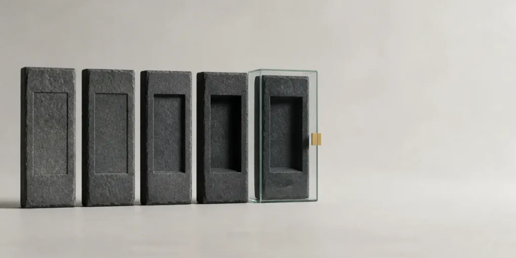 Five identical grey stone slabs in a row, each carved with the same blank panel but cut progressively deeper; the deepest-carved slab is sealed inside a glass case with a small brass clasp